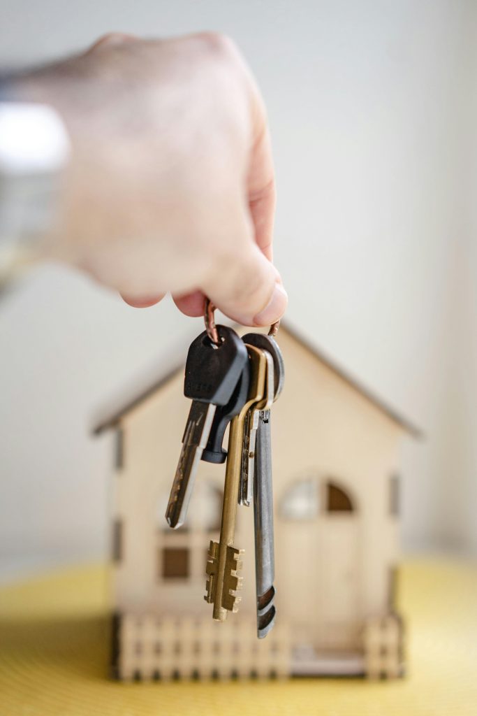 pexels photo 7599735 7599735 Close-up of a hand holding keys with a miniature wooden house in the background, symbolizing real estate investment.